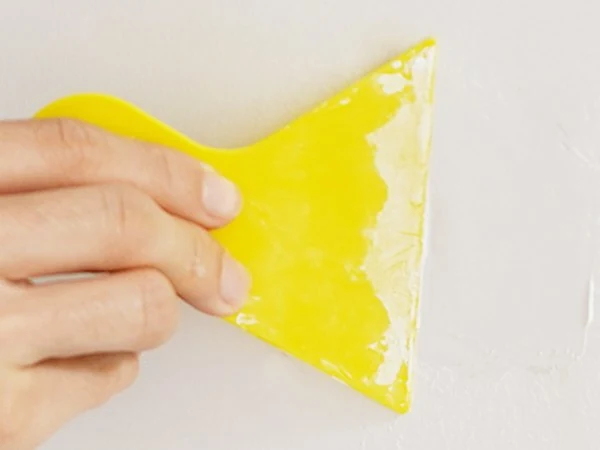 A yellow scraper smoothing repair paste over a wall